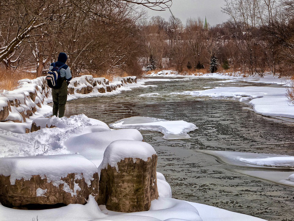 Person fishing with kids in cold weather by a snowy riverbank with trees in the background