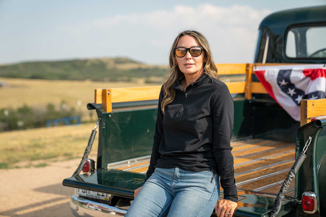 A woman wearing sunglasses, a black long-sleeve top, and blue jeans sits in the bed of a vintage green pickup truck outdoors with a blurred rural landscape in the background.