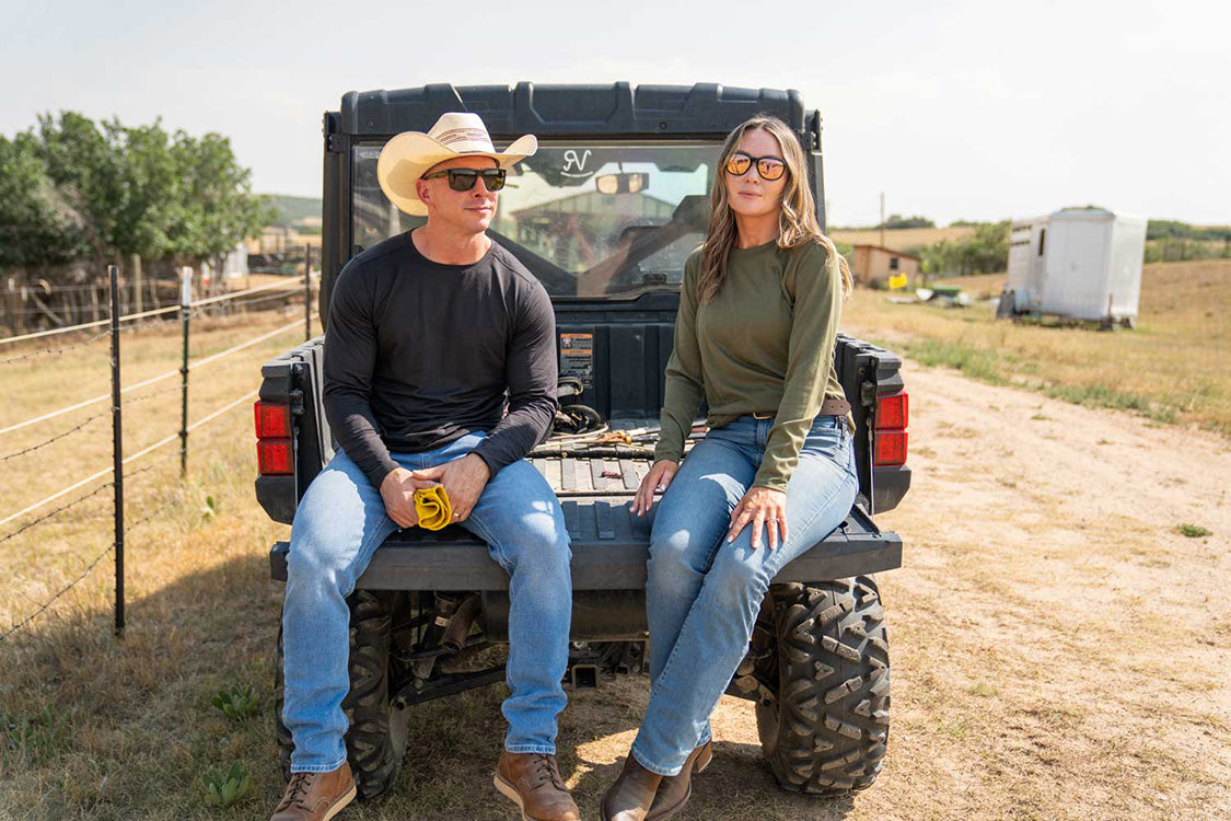 A man and a woman sit on the back of a black utility vehicle outdoors on a dirt road, with a fence, trees, and a small trailer in the background.