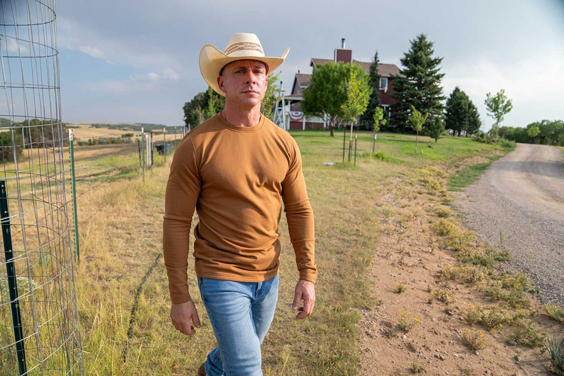 A man wearing a tan long-sleeve shirt, blue jeans, and a cowboy hat walking outdoors on a dirt road next to a wire fence, with a rural house and trees in the background.
