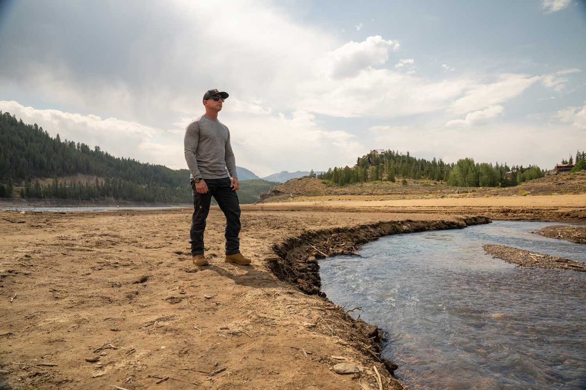 A man wearing a wool gray long-sleeve shirt, black pants, and brown boots stands on a dry, sandy riverbank next to a flowing stream, with mountains and trees in the background under a cloudy sky.